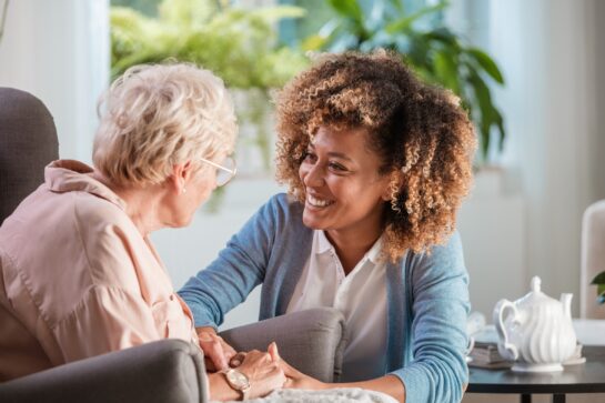 Lady with brown curly hair smiling at an older lady sitting in an arm chair