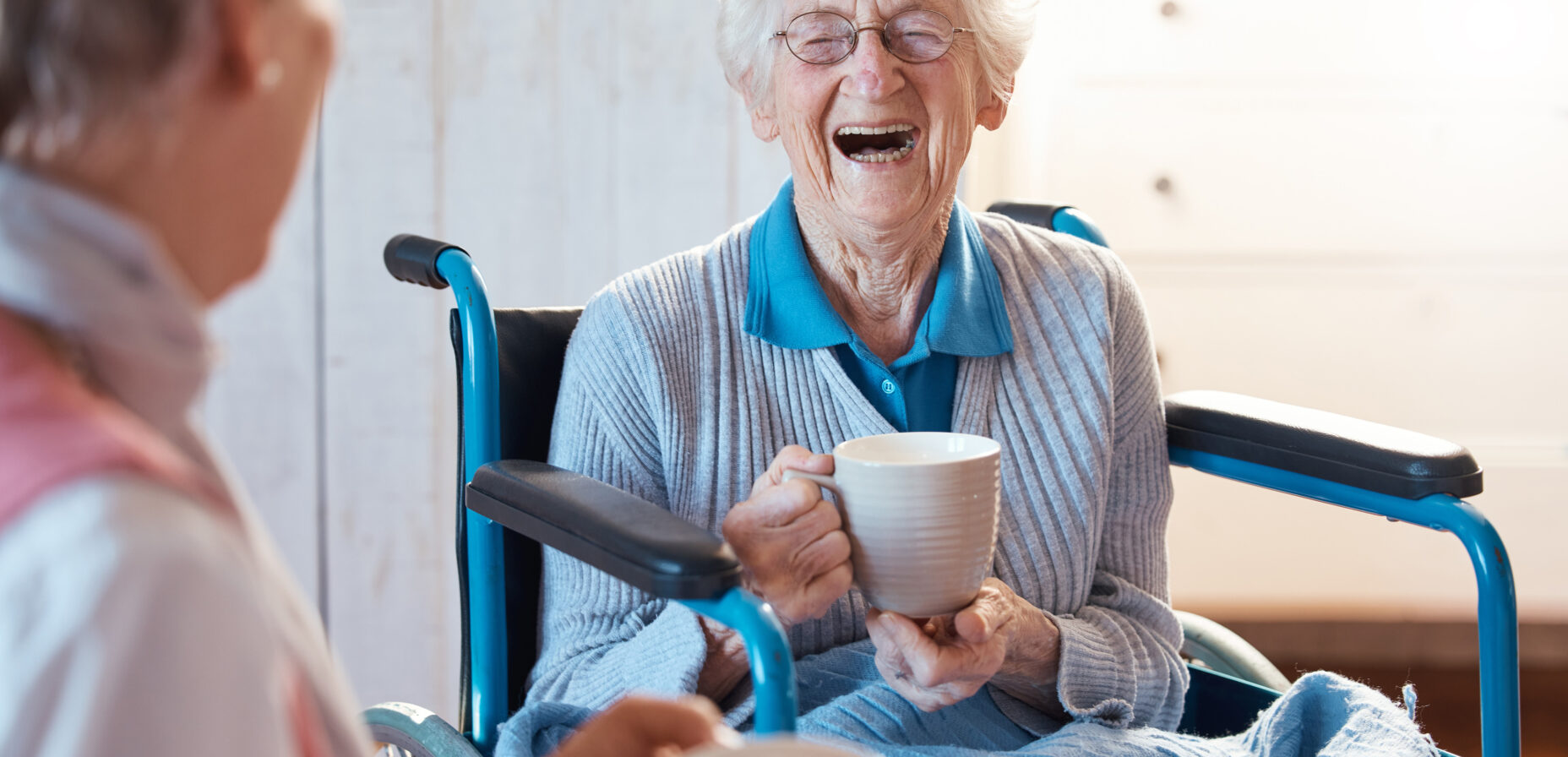 Older lady with grey hair sitting in a wheelchair with a cup of tea with a big smile on her face